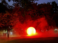 Eine leuchtend gelbe Kugel, umgeben von rotem Nebel, erhellt einen Park in der Abenddämmerung, in dessen Hintergrund Bäume und die Silhouetten von Menschen zu sehen sind.