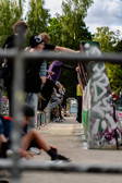 Eine Gruppe von Menschen versammelt sich an einem bewölkten Tag auf einer mit Graffiti bedeckten Skateparkrampe, einige sitzend, einige stehend, mit Bäumen und Grün im Hintergrund.