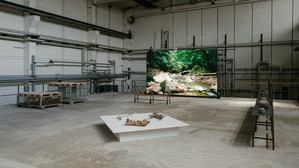 Auf dem Foto ist das Kunstwerk von Uriel Orlow zu sehen in einer großen Halle des ehemaligen Kohlekraftwerks in Chemnitz Nord. Das Kunstwerk besteht aus einer digitalen Leinwand und Elementen, die am Boden auf einer Platte liegen.