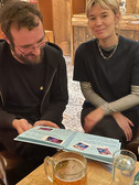 Ein Mann und eine Frau sitzen an einem Holztisch und schauen auf ein geöffnetes Fotoalbum. Der Mann lächelt und schaut auf das Album hinunter, während die Frau in die Kamera schaut und lächelt. Ein Glas Bier steht auf dem Tisch vor ihnen.