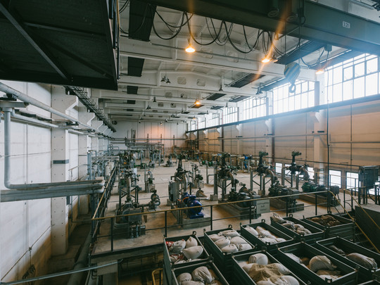 Eine weitläufige Industrieanlage mit hohen Decken, großen Deckenlampen und zahlreichen großen Rohren und Maschinen. Im Vordergrund sind mehrere große Behälter mit weißen Säcken zu sehen. Sonnenlicht fällt durch hohe Fenster auf der rechten Seite ein.