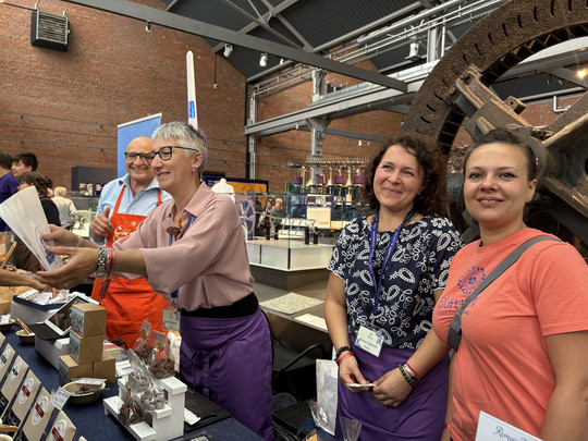 Vier lächelnde Menschen stehen hinter einem Tisch mit verschiedenen Pralinen und Leckereien auf einem Markt oder einer Veranstaltung in einer Halle, mit Industriemaschinen und Backsteinmauern im Hintergrund. Eine Frau reicht einem Kunden einen Artikel.
