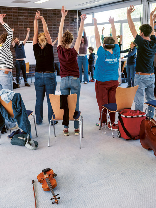 In einem hellen Raum mit großen Fenstern steht eine Gruppe von Menschen mit erhobenen Armen im Kreis. Stühle und Musikinstrumente, darunter eine Geige und ein Cello, sind im Raum verteilt.