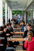 Die Menschen sitzen in Reihen an langen Tischen im Freien und spielen Schach unter einem überdachten Steg. Auf dem Platz herrscht reges Treiben mit Spielern und Zuschauern, und im Hintergrund sind Bäume zu sehen.