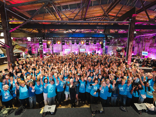 Eine große Gruppe von Menschen in passenden blauen Hemden steht in einem Innenraum, lächelt und winkt in die Kamera bei einer Veranstaltung unter einer Decke im industriellen Stil mit hellen Lichtern und einer Bühne im Hintergrund.