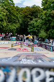 Ein Skateboarder führt einen Trick über einem Rail in einem mit Graffiti bedeckten Skatepark aus, während eine Zuschauermenge zusieht und im Hintergrund Bäume zu sehen sind.
