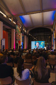 Ein Publikum sitzt in einem großen, farbenfrohen Saal und beobachtet einen Mann, der auf einer großen Leinwand im vorderen Bereich spricht. Der Raum ist mit farbenfrohen Bannern geschmückt, und die Atmosphäre wirkt förmlich und aufmerksam.