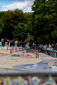 Ein Skateboarder führt einen Trick auf einem Rail in einem mit Graffiti bedeckten Skatepark aus, während im Hintergrund eine Zuschauermenge zusieht, mit Bäumen und einem teilweise bewölkten Himmel im Hintergrund.