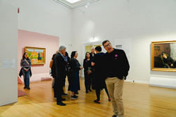 Eine Gruppe von Menschen steht in einer Kunstgalerie, einige betrachten die Gemälde an den Wänden. Ein Mann im Vordergrund lächelt und lehnt sich in Richtung der Kamera. Der Raum hat einen Holzboden und weiße und rosa Wände.