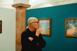 Ein glatzköpfiger Mann mit Brille und schwarzem Hemd steht in einer Kunstgalerie und betrachtet nachdenklich die farbenfrohen Gemälde an einer türkisfarbenen Wand.