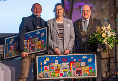 Drei formell gekleidete Personen stehen lächelnd in einem Haus. Zwei von ihnen halten bunte gerahmte Kunstwerke, die lebendige Stadtszenen darstellen; die dritte Person hält einen Blumenstrauß. Im Hintergrund sind ein Mikrofon und Bühnenscheinwerfer zu sehen.