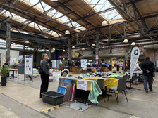 In einer großen Halle im Industriestil findet eine Kreativmesse statt, auf deren Tischen bunte Kunstwerke und Kunsthandwerk ausgestellt sind. Die Leute stöbern in den Ständen, und von der Decke hängen Dekorationen. Helles Licht fällt durch die Oberlichter im Holzdach.