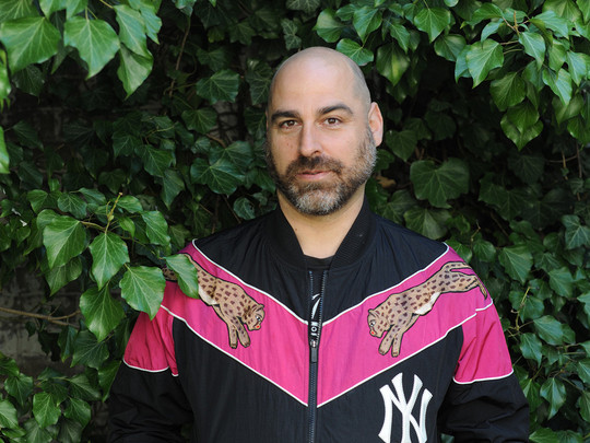 Eine Person mit Glatze und Bart steht vor einem grünen Hintergrund. Sie trägt eine schwarze Jacke mit rosa und magentafarbenen Akzenten, Gepardenmotiven und einem Logo der New York Yankees. Ihre Hände stecken in den Jackentaschen.