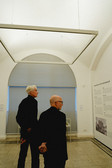 Zwei Männer mit kurzen Haaren und dunkler Kleidung stehen in einem minimalistischen Galerieraum und betrachten bei sanftem Tageslicht einen Ausstellungstext an einer weißen Wand.