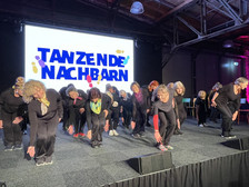 Eine Gruppe älterer Frauen in schwarzer Kleidung führt auf der Bühne einen Synchrontanz auf, wobei sie sich mit ausgestreckten Armen nach vorne beugen. Hinter ihnen erscheint auf einer Leinwand der Schriftzug "TANZENDE NACHBARN" auf einem bunten Hintergrund.
