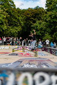 Ein Skateboarder balanciert auf einer Schiene in einem mit Graffiti bedeckten Skatepark, umgeben von einer Menge Schaulustiger und üppig grünen Bäumen im Hintergrund.
