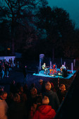 Eine kleine Band spielt nachts auf einer Freilichtbühne, umgeben von bunten Lichtern und Bäumen, während eine Menschenmenge im Vordergrund zusieht.