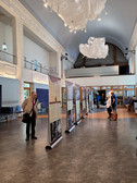 Ein Mann liest eine Ausstellungstafel in einer geräumigen Halle mit hohen Decken, moderner Beleuchtung und hängenden Stoffdekorationen. Im Hintergrund sind andere Menschen zu sehen, einige stehend, andere sitzend.