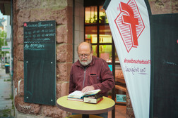Ein älterer Mann mit Brille und Bart steht an einem runden Tisch vor einem Gebäude und liest ein aufgeschlagenes Buch, neben dem sich weitere Bücher stapeln. In der Nähe befinden sich Kreidetafeln und eine weiße Fahne mit roter Schrift und einem Logo.