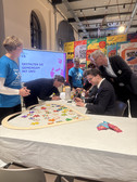 Eine Gruppe von Menschen arbeitet in einem bunt dekorierten Raum um einen Tisch mit einer großen herzförmigen Tafel und bunten Gummibärchen. Einige sitzen, schreiben oder ordnen Gegenstände, während andere in der Nähe stehen. Auf einem Bildschirm wird ein deutscher Text angezeigt.