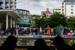 Bei einer städtischen Veranstaltung im Freien versammelt sich eine Menschenmenge an einem Teich und beobachtet einen Straßenkünstler, der Seifenblasen macht. Menschen aller Altersgruppen genießen die lebhafte Szene, die von modernen Gebäuden und Grünflächen umgeben ist.