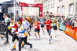 Eine Gruppe von Läufern in farbenfrohen Outfits nimmt an einem Stadtlauf teil, der unter einem roten Bogen mit der Aufschrift "START" beginnt, während Zuschauer die Kopfsteinpflasterstraße in einem historischen Stadtviertel säumen.