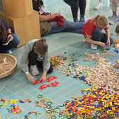 Kinder sitzen und knien auf einem blau gekachelten Boden und sortieren und ordnen bunte geometrische Formen und Holzteile zu Mustern an. Erwachsene und weitere Kinder stehen in der Nähe und schauen zu oder nehmen an der Aktivität teil.