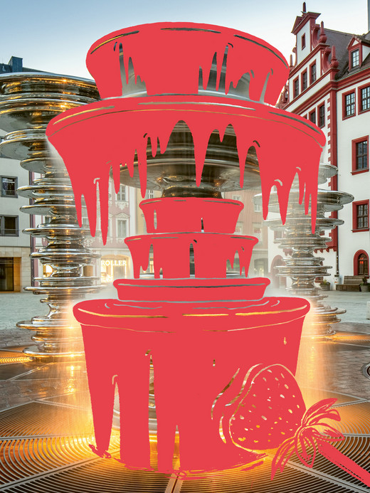 Ein digital bearbeitetes Bild zeigt einen Stadtbrunnen mit einer Grafiküberlagerung, die einem Schokoladenbrunnen mit roter tropfender Schokolade und einer Erdbeere ähnelt. Im Hintergrund sind historische Gebäude auf einem Kopfsteinpflasterplatz zu sehen.