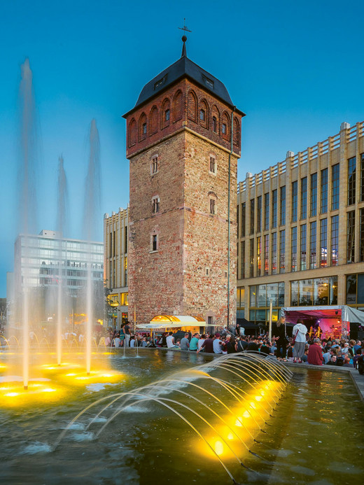 Eine geschäftige Abendszene mit Menschen, die sich um einen großen, beleuchteten Brunnen versammelt haben. In der Nähe steht ein historischer Backsteinturm, neben einem modernen Glasgebäude. Der Himmel ist klar und trägt zur lebhaften Atmosphäre bei.
