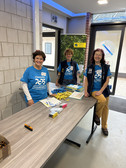 Drei lächelnde Freiwillige in blauen T-Shirts stehen hinter einem Anmeldetisch mit Ausweisen, Stiften und Flyern in einem Foyer. Im Hintergrund sind ein Transparent und Glastüren zu sehen.