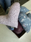 Eine Schachtel gefüllt mit herzförmigen Kissen aus verschieden gemusterten Stoffen, darunter lila mit Leopardenmuster, blau mit Blumenmuster und kastanienbraun mit Paisleymuster, auf einem hellen Tisch.