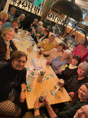 Eine Gruppe lächelnder Frauen versammelt sich um einen langen Holztisch in einem gemütlichen Restaurant, hält Getränke in der Hand und schaut in die Kamera. Speisekarten, Getränke und Tischgegenstände sind zu sehen; ein großes Fahrrad ziert die Wand im Hintergrund.