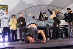 Ein Breakdancer balanciert auf einer Hand mit ausgestreckten Beinen in der Luft und tritt auf der Bühne auf, während im Hintergrund eine Gruppe von Kindern zuschaut und jubelt. Die Szene ist energiegeladen und lebendig.