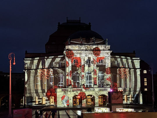 Ein verziertes Gebäude wird nachts mit einer farbenfrohen Projektion aus rotem Mohn und abstrakten Formen auf seiner Fassade beleuchtet, die sich lebhaft vom dunklen Himmel abhebt.