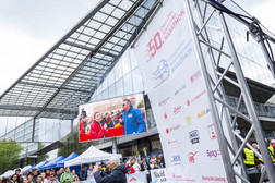 Eine große Marathonveranstaltung im Freien mit einer Menschenmenge, Sponsorenbannern und einem großen Bildschirm, auf dem zwei Personen sprechen. Die Szene spielt sich an einem bewölkten Tag vor einem modernen Glasgebäude ab.
