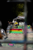 Ein Skateboarder in einem gelben Kapuzenpulli vollführt einen Trick auf einer bemalten Treppe in einem Skatepark, beobachtet von Zuschauern. Die Rampen sind mit Graffiti verziert und im Hintergrund ist ein Zelt mit Bannern zu sehen.