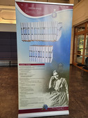 Eine vertikale Schautafel über Edith Stein zeigt ein großes Foto von ihr im Sitzen, eine Sammlung von Buchrücken im oberen Bereich, einen Text mit einer Beschreibung ihrer Werke und eine dekorative Blumengrafik im unteren Bereich.