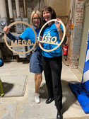 Zwei Frauen stehen in einem Innenraum auf einem Betonboden, lächeln und halten runde Holzschilder mit der Aufschrift "MEGA" und "LABO". Beide tragen blaue Hemden; eine trägt ein Jeanskleid mit Brille, die andere eine schwarze Hose. Hinter ihnen befinden sich freiliegende Rohre und Backsteinwände.