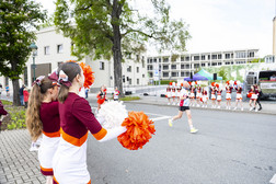 Cheerleader in kastanienbraunen und weißen Uniformen mit orangefarbenen Pompons stehen auf einem Bürgersteig und feuern einen Läufer auf der Straße an, während sich im Hintergrund auf der anderen Straßenseite weitere Cheerleader versammeln.