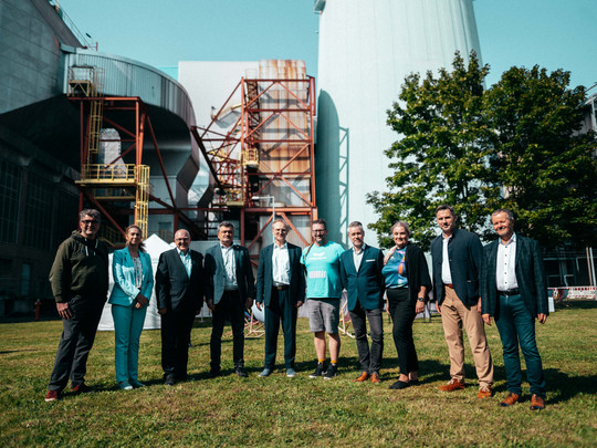 Eine Gruppe von zehn Personen steht draußen auf einer Rasenfläche vor einer industriellen Kulisse. Die Gruppe besteht aus Männern und Frauen in Business- und Freizeitkleidung. Sie posieren in einer Reihe und lächeln in die Kamera, mit einem sonnigen Himmel und Bäumen im Hintergrund.