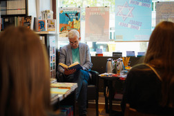 Ein älterer Mann mit grauem Haar und Brille sitzt in einer Buchhandlung und liest einem Publikum aus einem Buch vor. Um ihn herum sind Bücherregale, Poster und ein Tisch mit Schreibwaren zu sehen. Das Foto ist von hinten auf zwei Zuhörer aufgenommen.