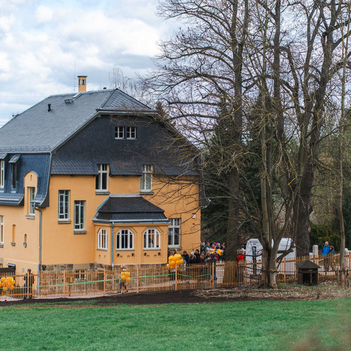 Ein großes gelbes Haus mit dunklem Dach ist von kahlen Bäumen und einer Wiese umgeben. Gelbe Luftballons schmücken den umzäunten Hof, und mehrere Menschen haben sich in der Nähe des Eingangs versammelt. Der Himmel ist teilweise bewölkt.