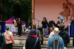 Eine Band spielt auf einer Bühne im Freien, während Menschen in Jacken und Kapuzenpullovern zusehen. Der Bühnenhintergrund zeigt eine Illustration von tanzenden Figuren, und im Hintergrund sind Bäume zu sehen.