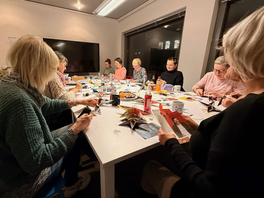 Eine Gruppe von Menschen sitzt in einem gut beleuchteten Raum um einen mit Bastelmaterial, Zeitschriften und Scheren gedeckten Tisch und beschäftigt sich mit Bastelarbeiten.
