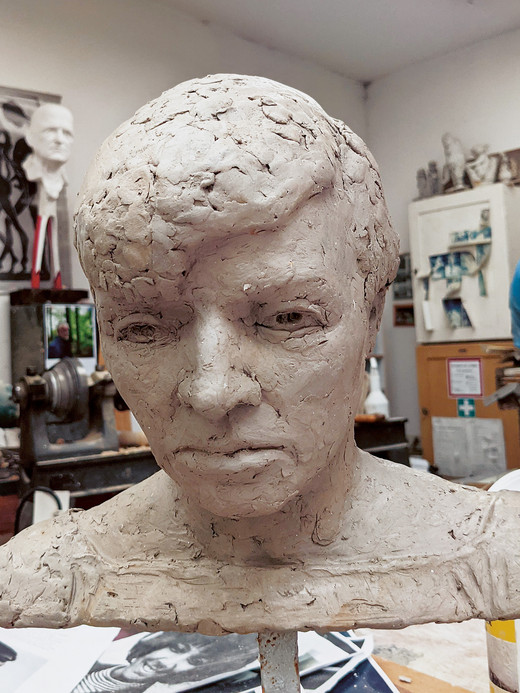 Eine Tonbüste eines menschlichen Kopfes mit detaillierten Gesichtszügen und strukturiertem Haar steht auf einem Tisch in einem Kunstatelier. Im Hintergrund sind verschiedene Werkzeuge, Kunstwerke und Skulpturen zu sehen.