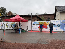 An einem nassen, bewölkten Tag malen Menschen bunte Graffiti-Kunstwerke auf eine weiße Barrikade unter einem roten Zeltdach, mit Backsteinbauten im Hintergrund.