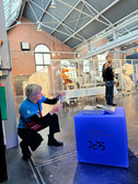 Eine Person geht in die Hocke und macht ein Foto mit einem Smartphone in einer Ausstellungshalle im Industriestil mit Glasvitrinen und Schaufensterpuppen in Schutzanzügen. Im Vordergrund steht ein blauer Würfel mit der Aufschrift "Chemnitz Kulturhauptstadt Europas 2025".