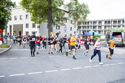 Eine Gruppe von Läufern in Sportkleidung nimmt an einem Straßenrennen auf einer Stadtstraße teil. Im Hintergrund sind Gebäude, Bäume und ein Zelt zu sehen. Die Zuschauer schauen vom Bürgersteig aus zu.