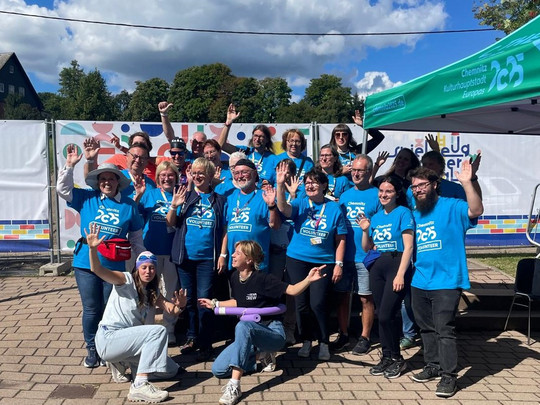 Eine Gruppe von Menschen in passenden blauen Hemden posiert und winkt unter einem sonnigen Himmel bei einer Veranstaltung im Freien. Sie stehen in der Nähe eines Zeltes mit der Aufschrift "25" und bunten Dekorationen im Hintergrund.