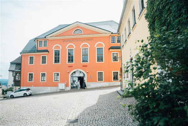 Am Ende einer Kopfsteinpflasterstraße steht ein historisches Gebäude mit einer leuchtend orangefarbenen Fassade. Der Eingang verfügt über einen gewölbten Durchgang und in seiner Nähe stehen ein paar Leute. In der Nähe parkt ein Auto. Auf der rechten Seite ist teilweise Grün zu sehen.
